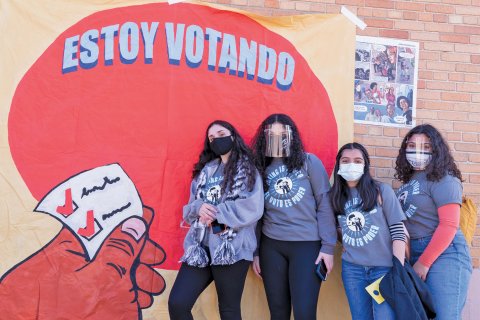 Students activists with the group Youth Empowered in the Struggle (YES) wear masks while standing in front of a sign that says, "Estoy Votando." 